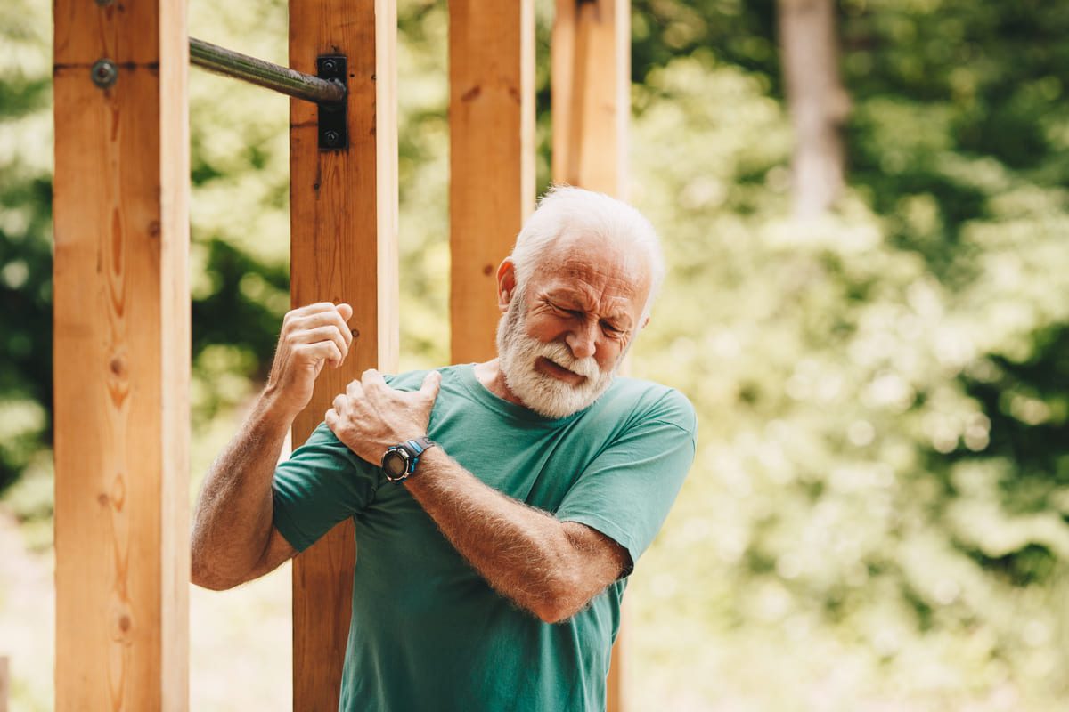 Älterer fitter Mann vor Fitnessgerät hält sich die Hand mit schmerzverzerrtem Gesicht an seine Schulter