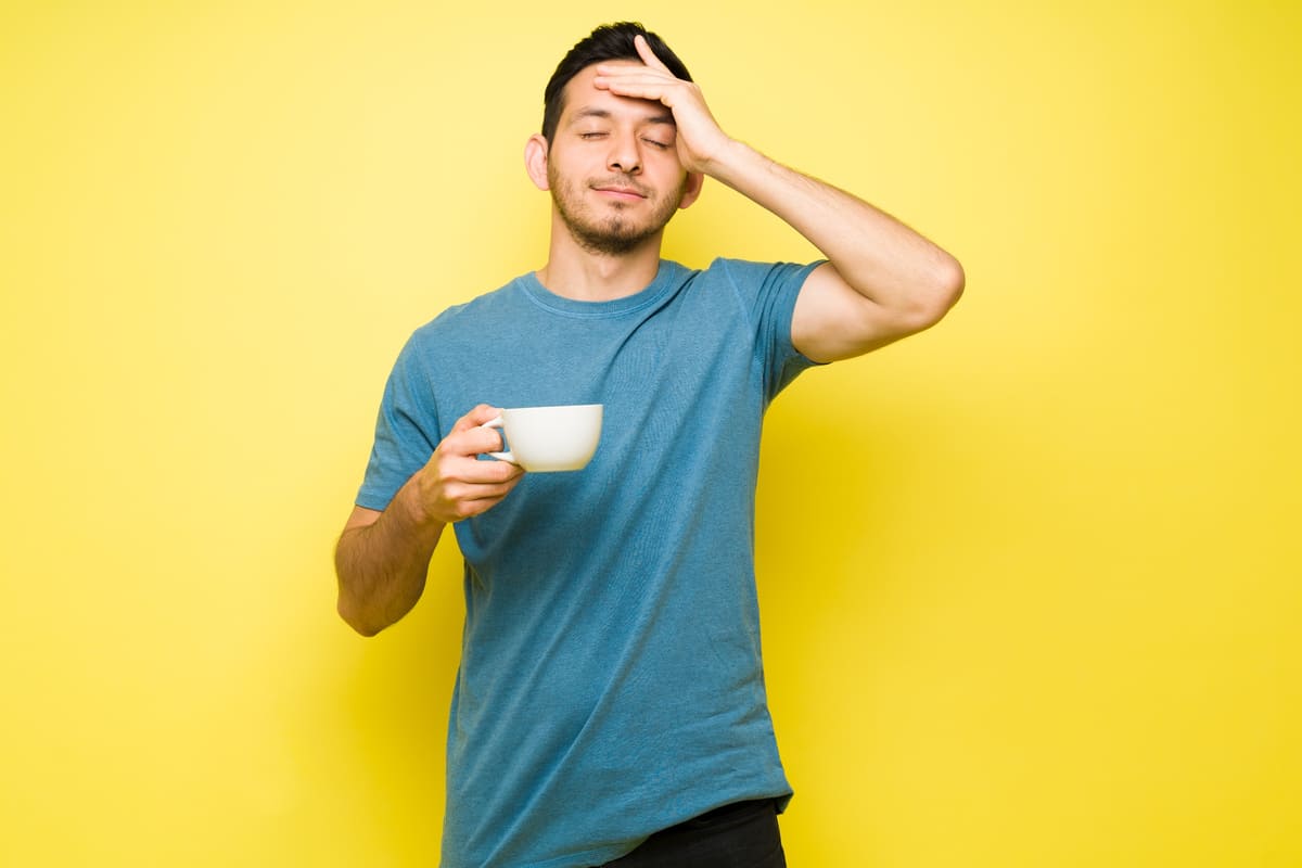 Mann vor gelbem Hintergrund mit Kaffee in der Hand fasst sich an den Kopf