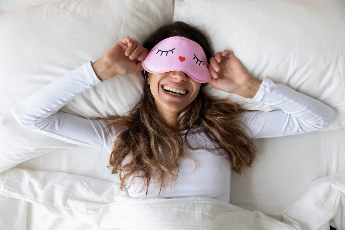 Frau im Bett liegend mit rosaroter Schlafmaske und lacht
